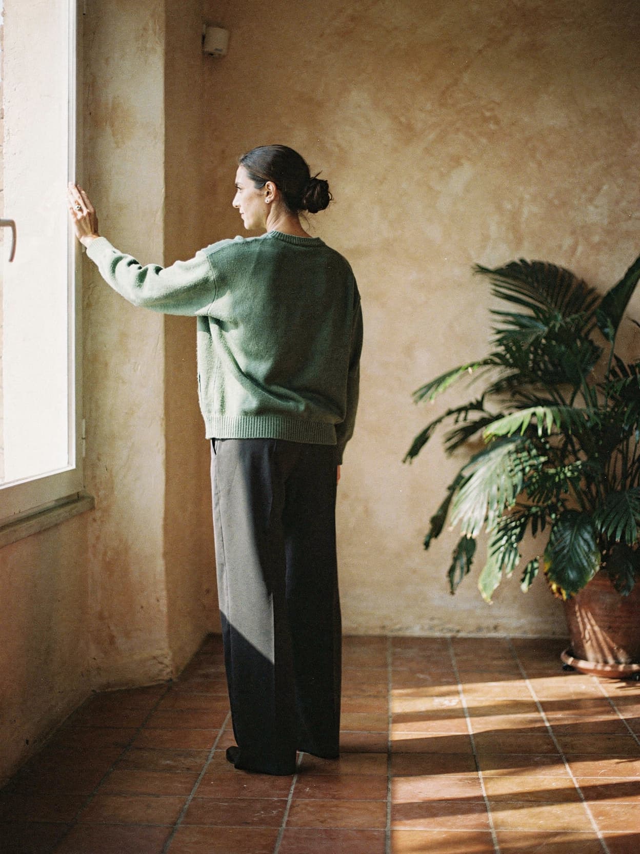 Marisol standing at a sunlit window, seen from behind
